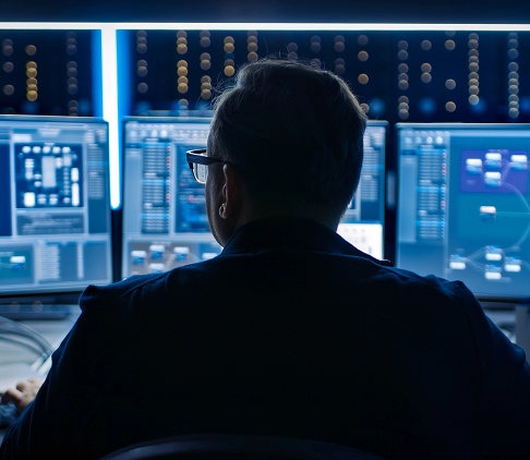 A man sits in a dark room surrounded by multiple glowing computer screens.