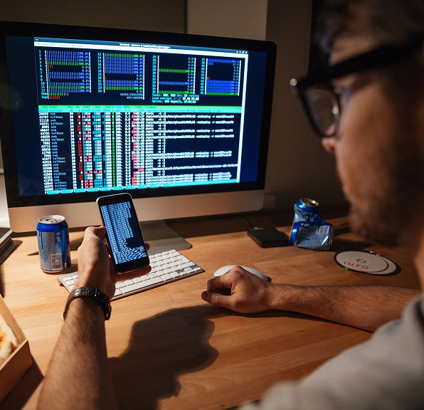 A man looks at a computer screen while holding his phone in his hand, engaged in a digital task.