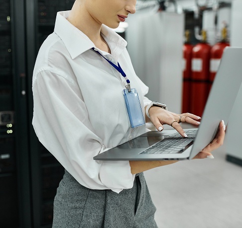 A woman in a white shirt and gray pants holds a laptop, looking focused and engaged in her work.