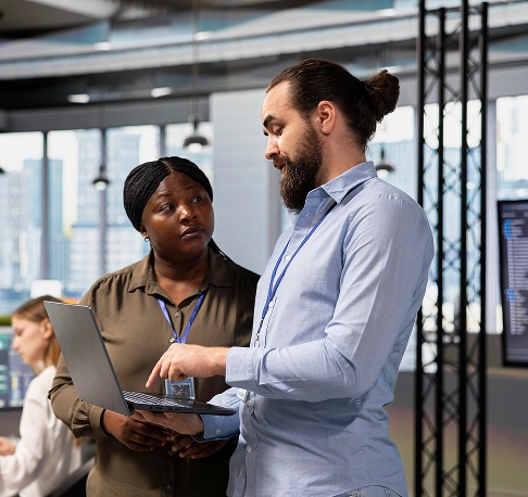 Two people standing together, looking at a laptop screen, engaged in discussion or collaboration.