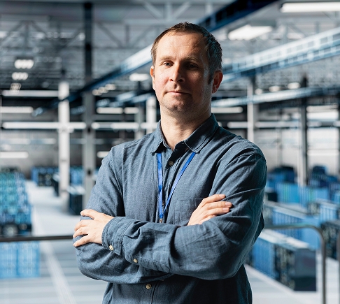 A man stands with arms crossed in a spacious warehouse, surrounded by shelves and industrial equipment.