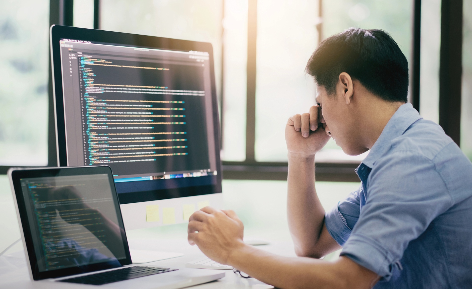 A man sitting at a desk, working on a laptop with a computer screen in front of him.