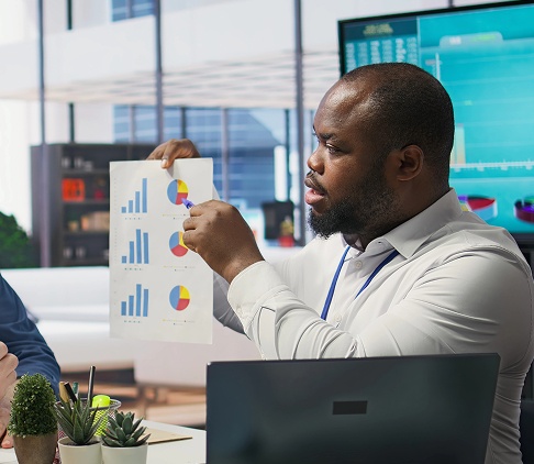Two men in an office analyzing graphs on a computer screen, discussing data insights and trends.