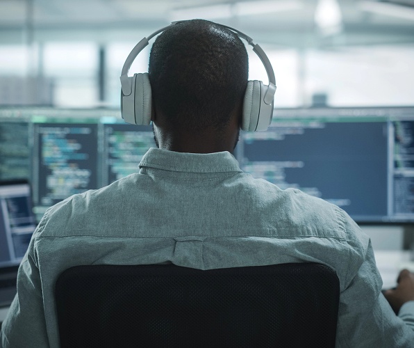 A man with headphones sits at a computer desk, focused on two monitors displaying various content.